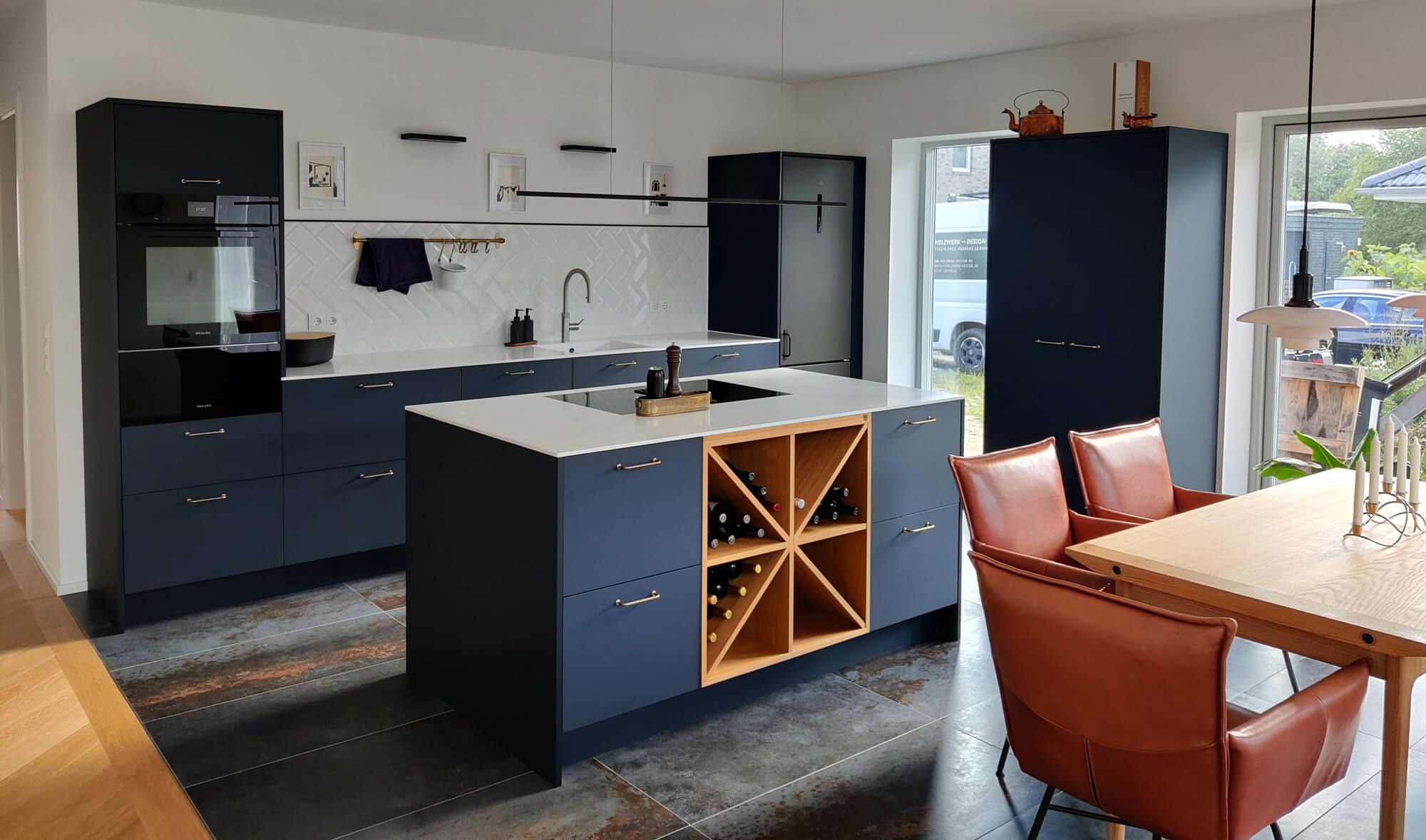 Eine moderne Küche mit marineblauen Schränken, einer weißen Arbeitsplatte, einem Weinkühlschrank und stilvollen Lederstühlen um einen Esstisch aus Holz.